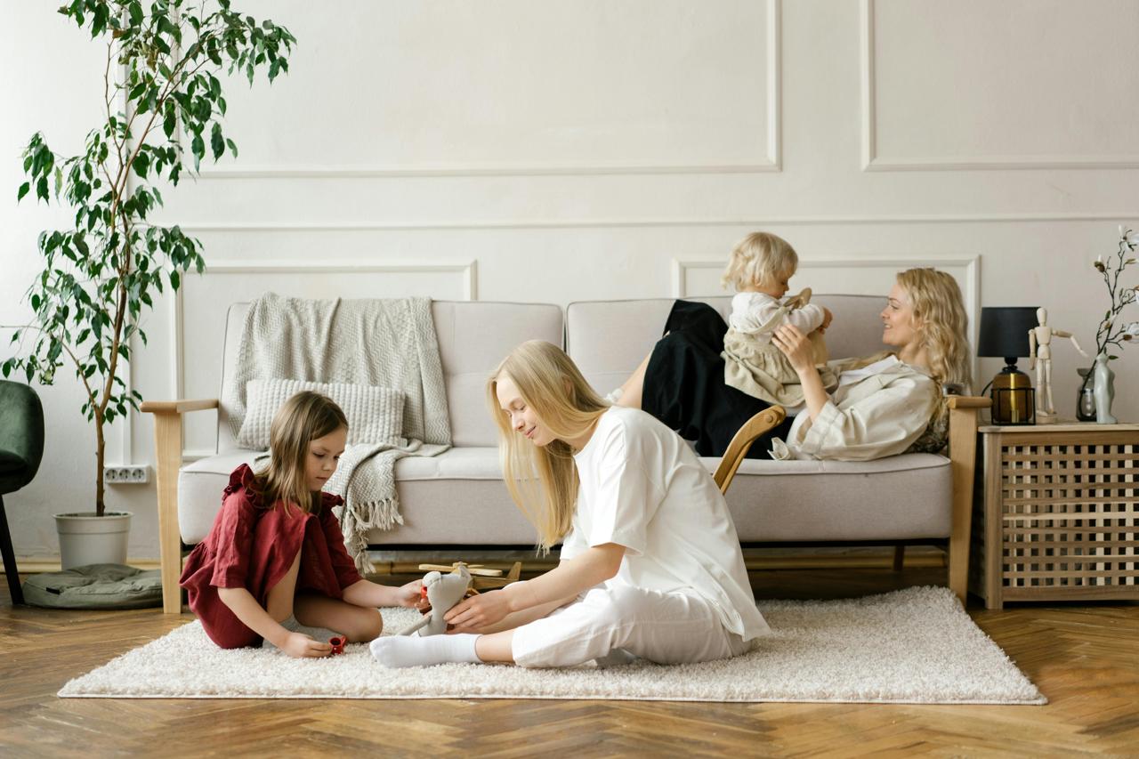 Three women and two children in a cozy living room setting—an older woman plays with a child on the couch, while two younger women and a girl sit on the rug playing together, reflecting warmth, connection, and multigenerational care.