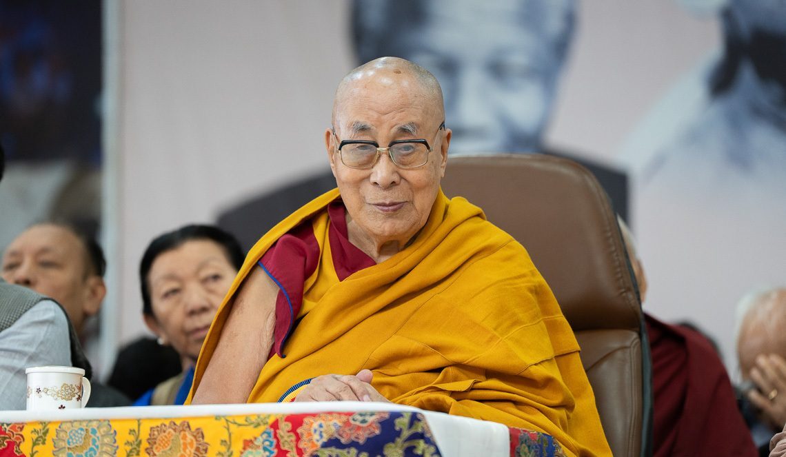The Dalai Lama giving a discourse, wearing saffron robes, some devotees behind him