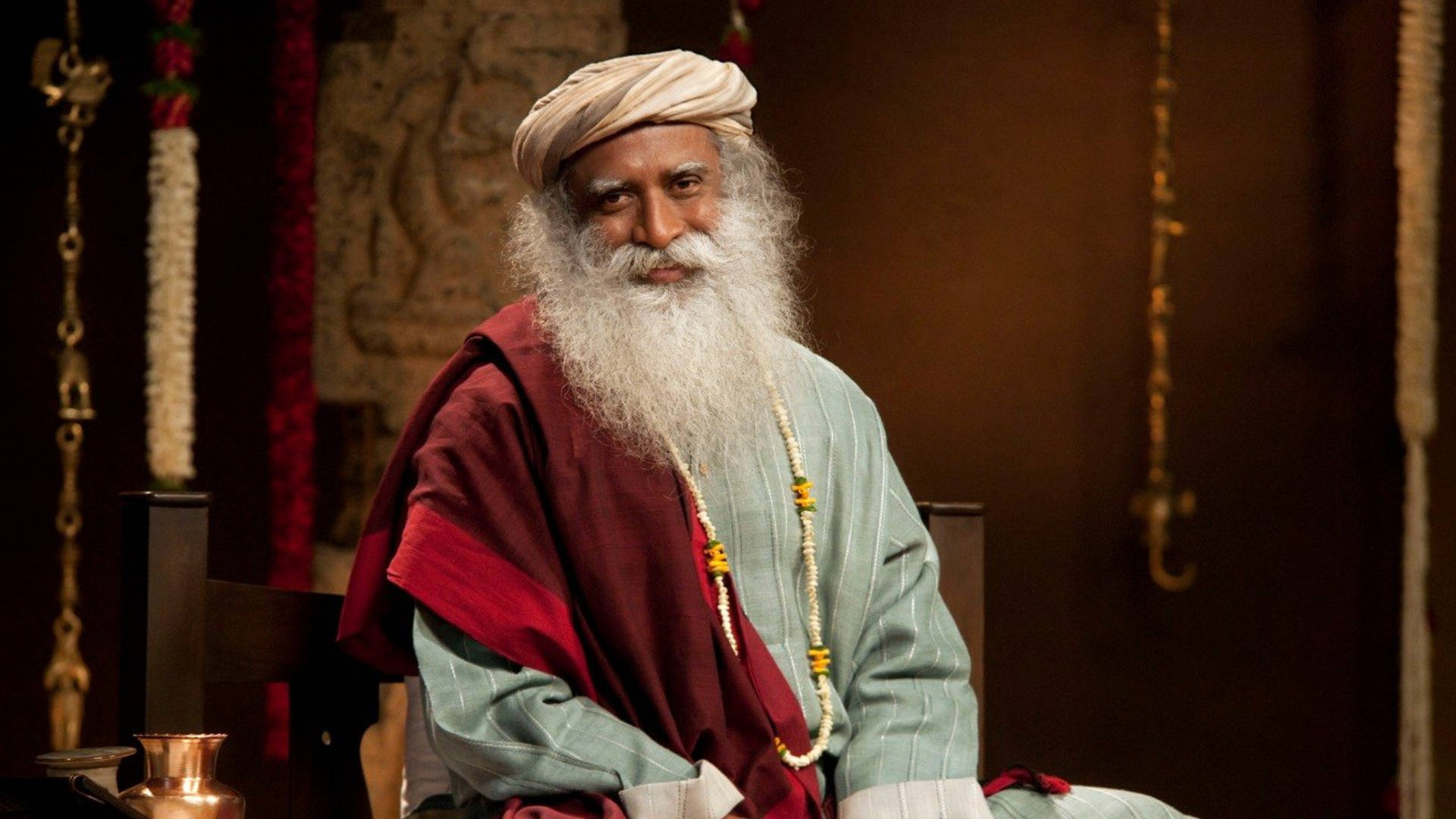 An elderly spiritual teacher with a long white beard and turban, seated indoors in traditional attire, gesturing gently while speaking.