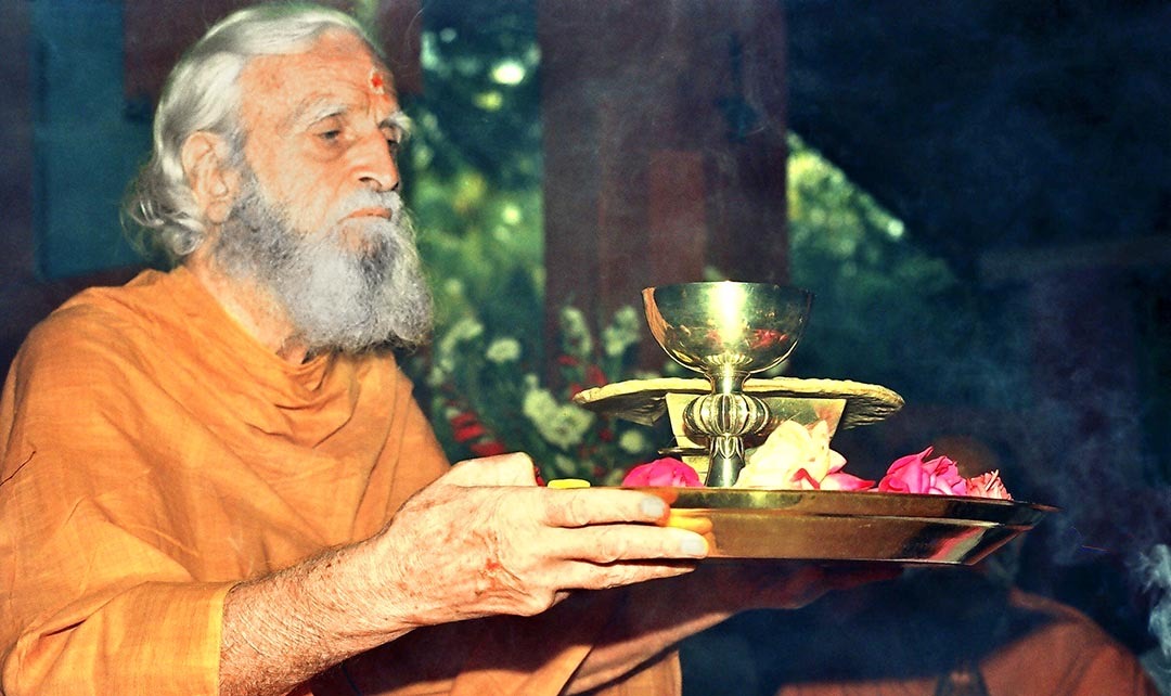 Bede Griffiths seated outdoors at Shantivanam Ashram in India, wearing saffron robes and speaking thoughtfully during a discussion on interfaith spirituality.