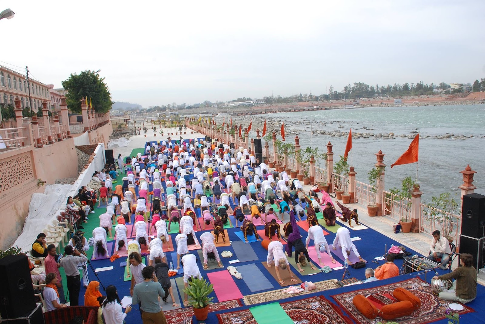 Hundreds of yoga practitioners performing Surya Namaskar at the International Yoga Festival at Parmarth Niketan Ashram in Rishikesh, beside the River Ganga with saffron flags and riverside promenade visible.