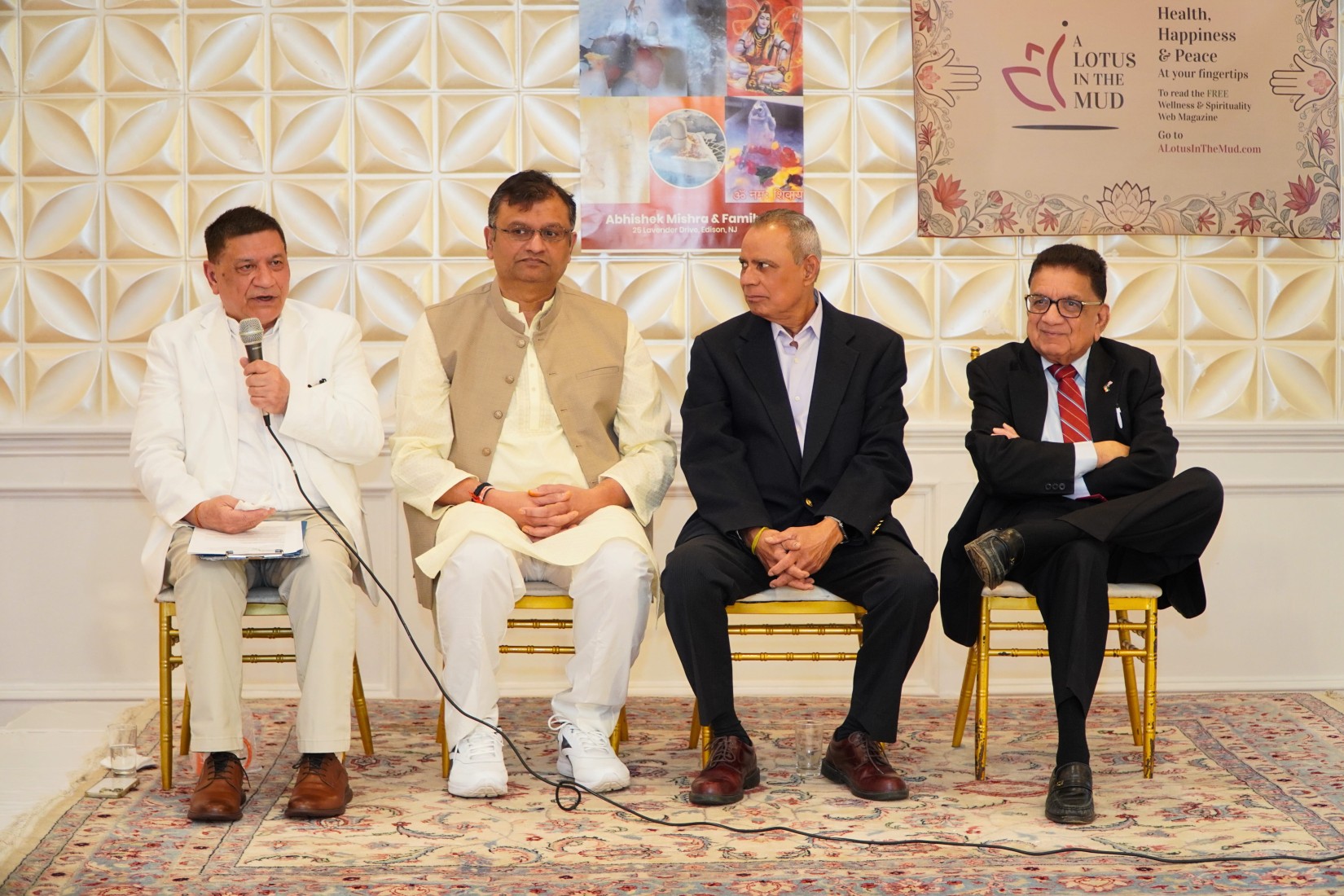 Parveen Chopra, Abhishek Mishra, Upendra Chivukula, and Peter (Pradip) Kothari sit side by side on stage, in that order from left to right. Seated on chairs, Parveen speaks into a handheld microphone while the others face the audience, listening attentively.